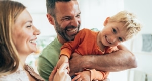 Eine Mutter und ein Vater haben ihren Sohn auf dem Arm und lachen.