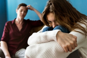 Eine junge Frau sitzt auf dem Sofa und hat die Arme verschränkt. Ihre Mutter schaut sie traurig an.