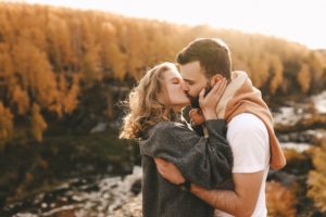 Ein Mann und eine Frau stehen vor einem herbstlichen Wald. Sie umarmen und küssen sich.