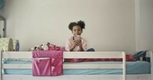 Ein Mädchen sitzt auf ihrem Hochbett und lächelt. Sie hält ein Handy in der Handy.