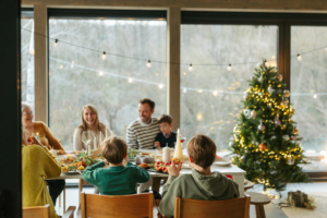 Eine Familie sitzt gemeinsam am Tisch. Die Leute lachen und reden, im Hintergrund steht ein Weihnachtsbaum.