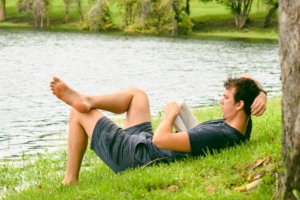 Ein Jugendlicher in kurzer Hose und T-Shirt liegt an einem See im Gras und liest ein Buch.