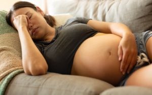 Eine schwangere Frau liegt auf einem Sofa. Sie hat die Augen geschlossen und hält sich die Hand vor den Kopf.