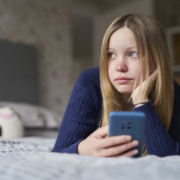 Teenager-Mädchen liegt einsam und traurig mit dem Handy auf dem Bett.