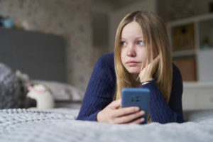 Teenager-Mädchen liegt einsam und traurig mit dem Handy auf dem Bett.