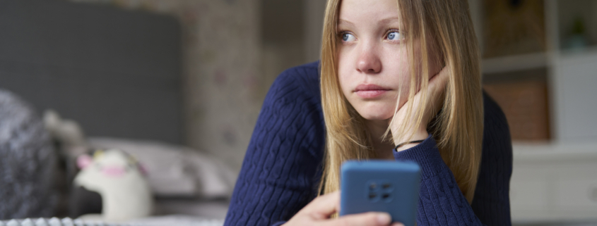Teenager-Mädchen liegt einsam und traurig mit dem Handy auf dem Bett.