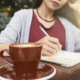 Eine Frau sitzt mit einem Cappuchino am Tisch und schreibt auf einem Block.